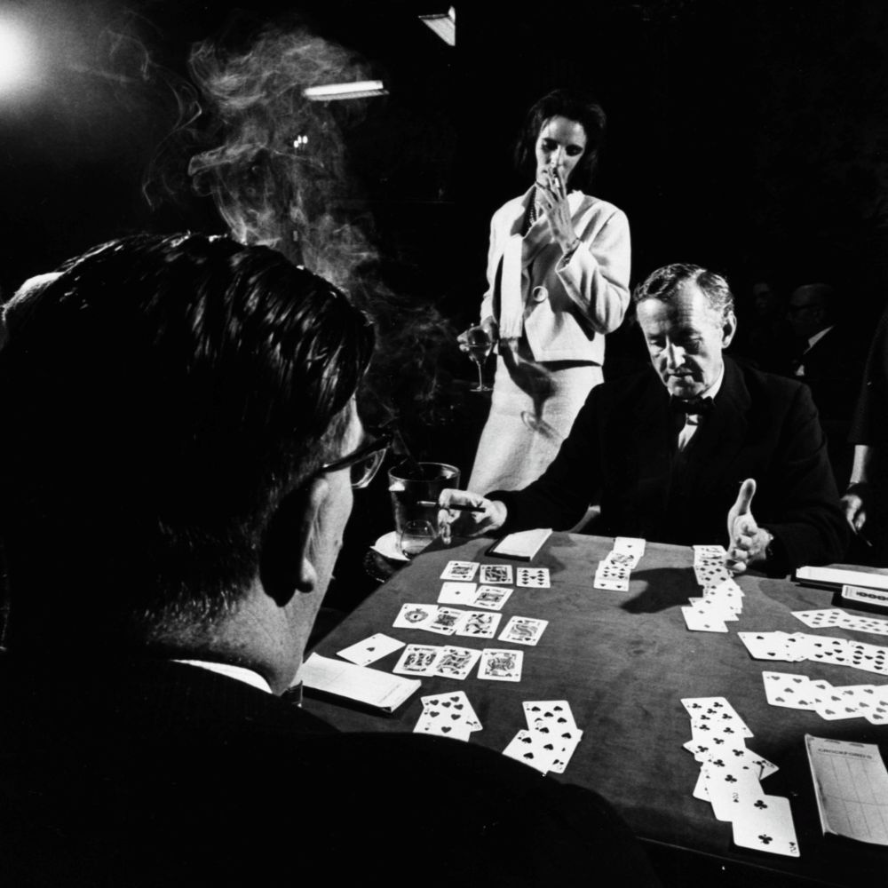 A black and white photograph of Ian Fleming playing bridge with a man. A woman is standing behind him watching. He is also holding a cigarette.