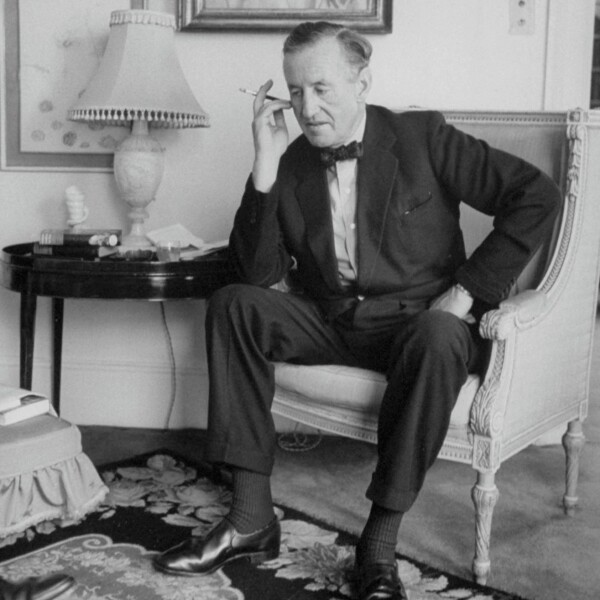 A black and white photograph of Ian Fleming seated in an armchair, wearing a suit and bow tie, holding a cigarette. A side table with a lamp and books is beside him, in a study-like interior.