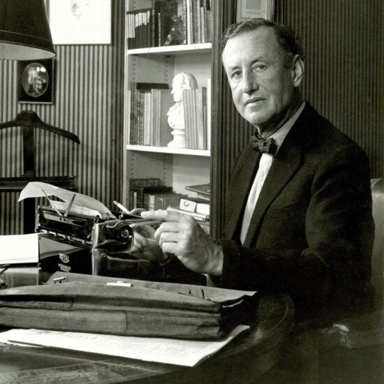 Black and white photograph of Ian Fleming sitting at a desk with a typewriter on it. He is looking at the camera.
