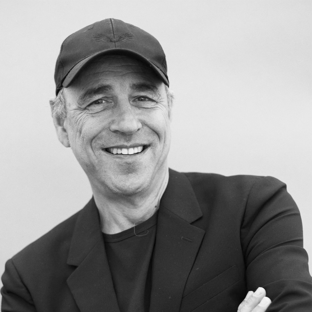 Black and white photograph of author Anthony Horowitz. He is a smiling middle aged white man wearing a suit jacket and baseball hat.