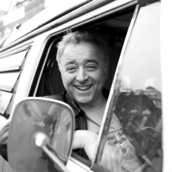 Black and white photograph of author Frank Cottrell Boyce, a smiling white man pictured driving a campervan.