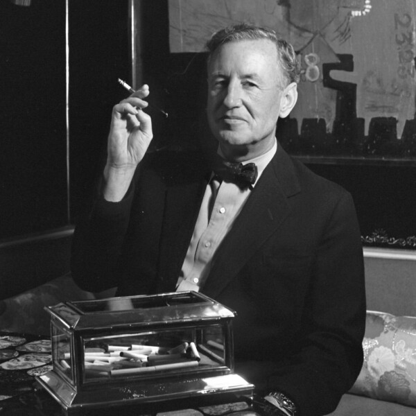 A black and white headshot of Ian Fleming sitting at a desk, wearing a suit and bow tie, holding a cigarette, smiling.