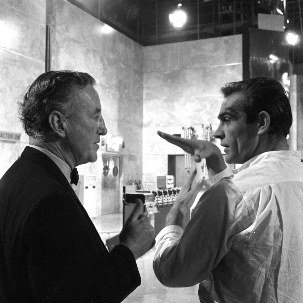 A black and white photograph of Ian Fleming in conversation with Sean Connery on a film set. The photograph is taken from behind so their profiles are in view.