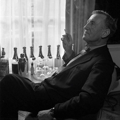 Black and white photograph of Ian Fleming seated indoors, holding a cigarette. Behind him is a table with bottles, glasses and a cocktail shaker.