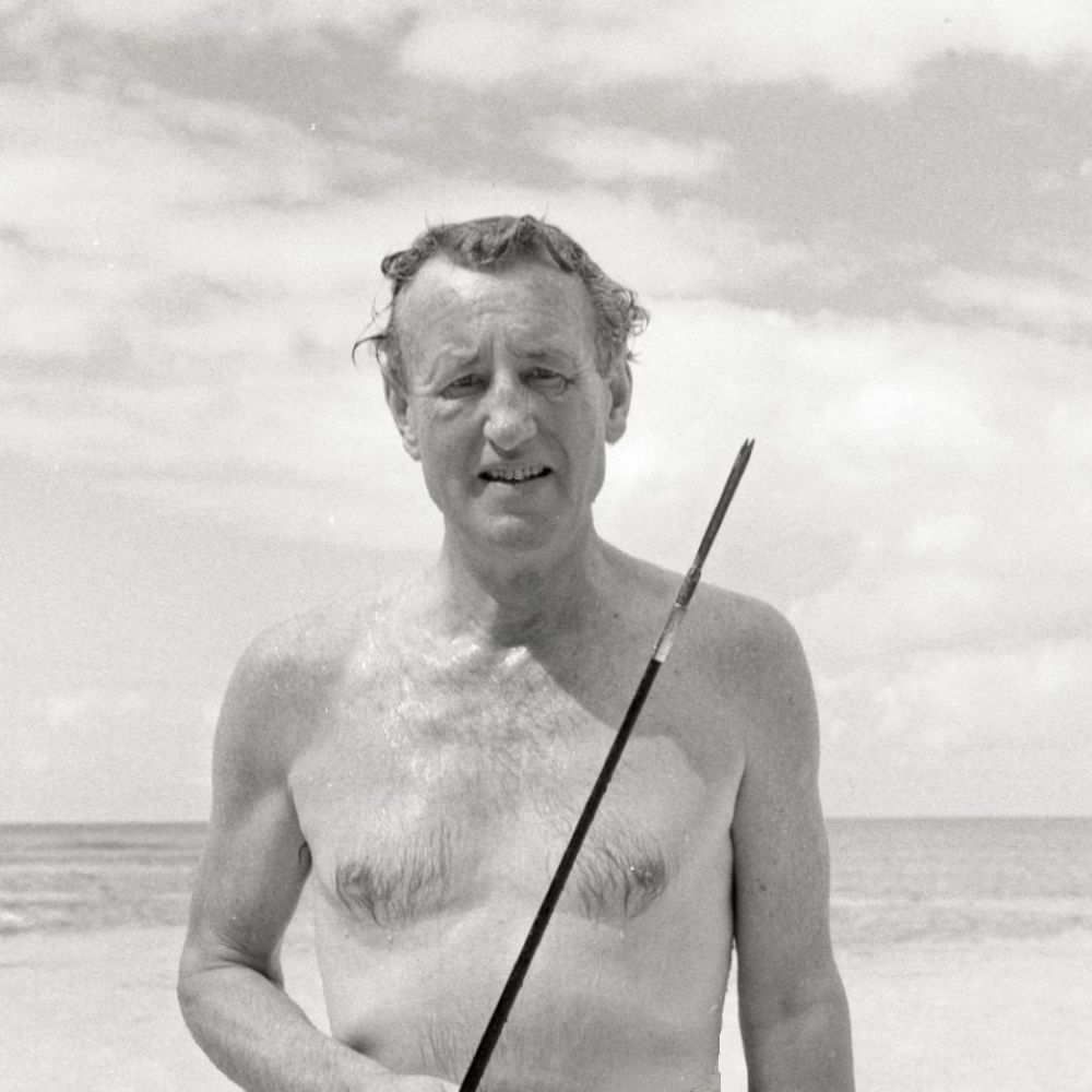 A black and white photograph of Ian Fleming with the sea in the background. He appears to have just come out of the sea and is holding a spear fishing pole.