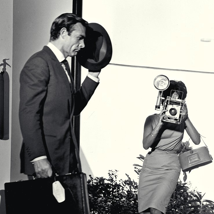 A black and white shot from the film Dr. No with a man, James Bond, shielding his face from a woman taking a photo of him as he walks past with a briefcase